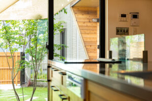 A modern kitchen with wooden cabinetry and a large window overlooking a green garden, creating an open and refreshing atmosphere.